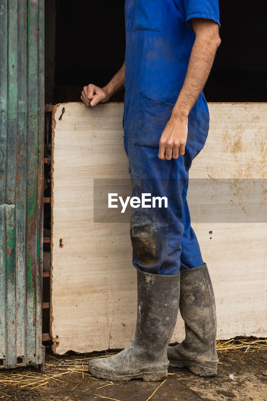 Cropped unrecognizable male farmer in blue uniform and galoshes standing in shabby farm building during work in rural area