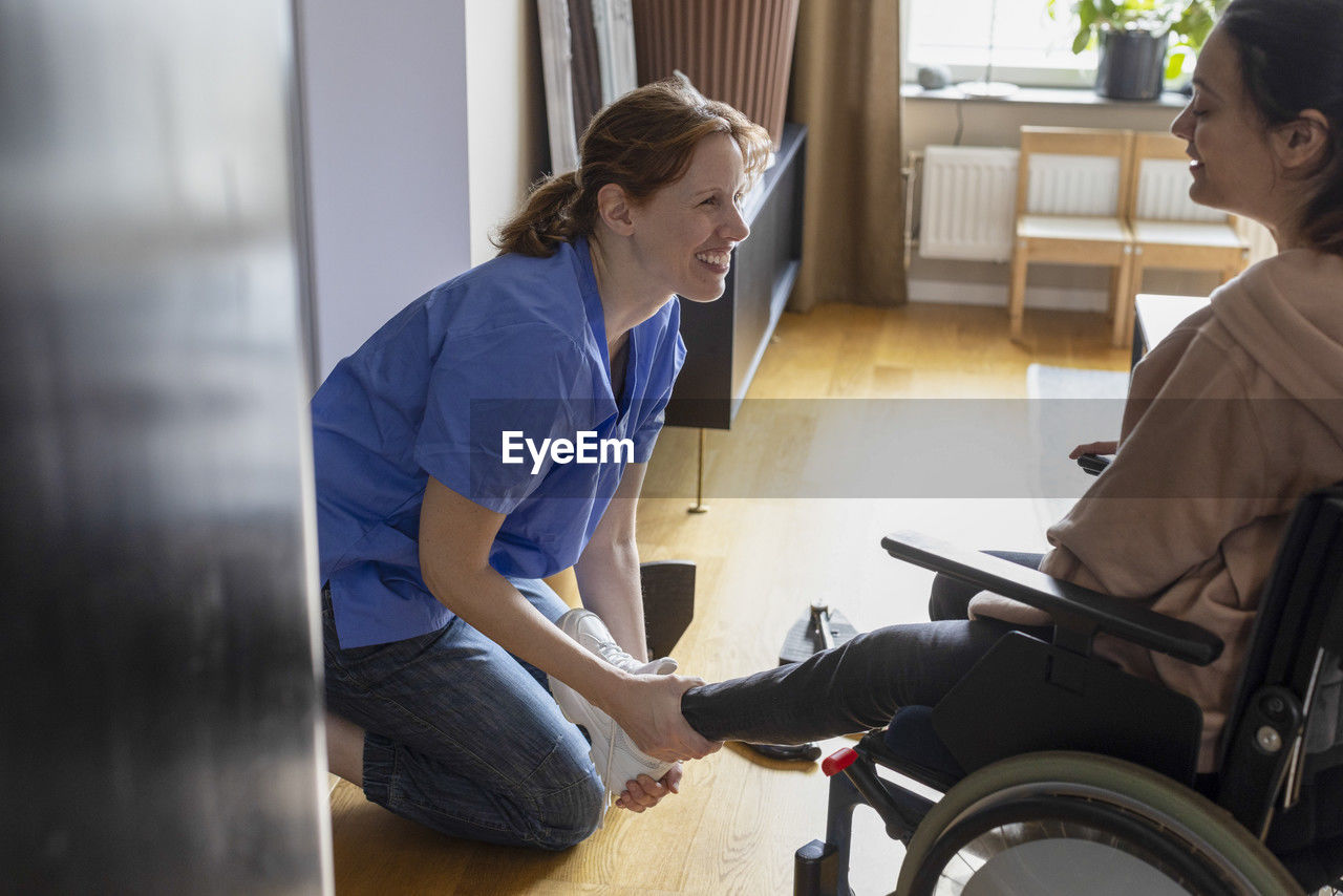 Happy nurse assisting woman with paraplegia while wearing shoes at home