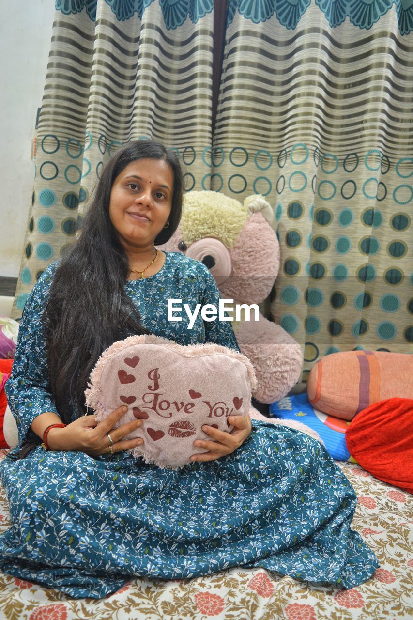 one person, women, indoors, adult, art, sitting, smiling, portrait, looking at camera, pattern, textile, front view, human face, lifestyles, female, happiness, furniture, emotion, person, childhood, clothing, child, home interior, blue, toy, full length, hairstyle, bed, casual clothing, domestic room, relaxation