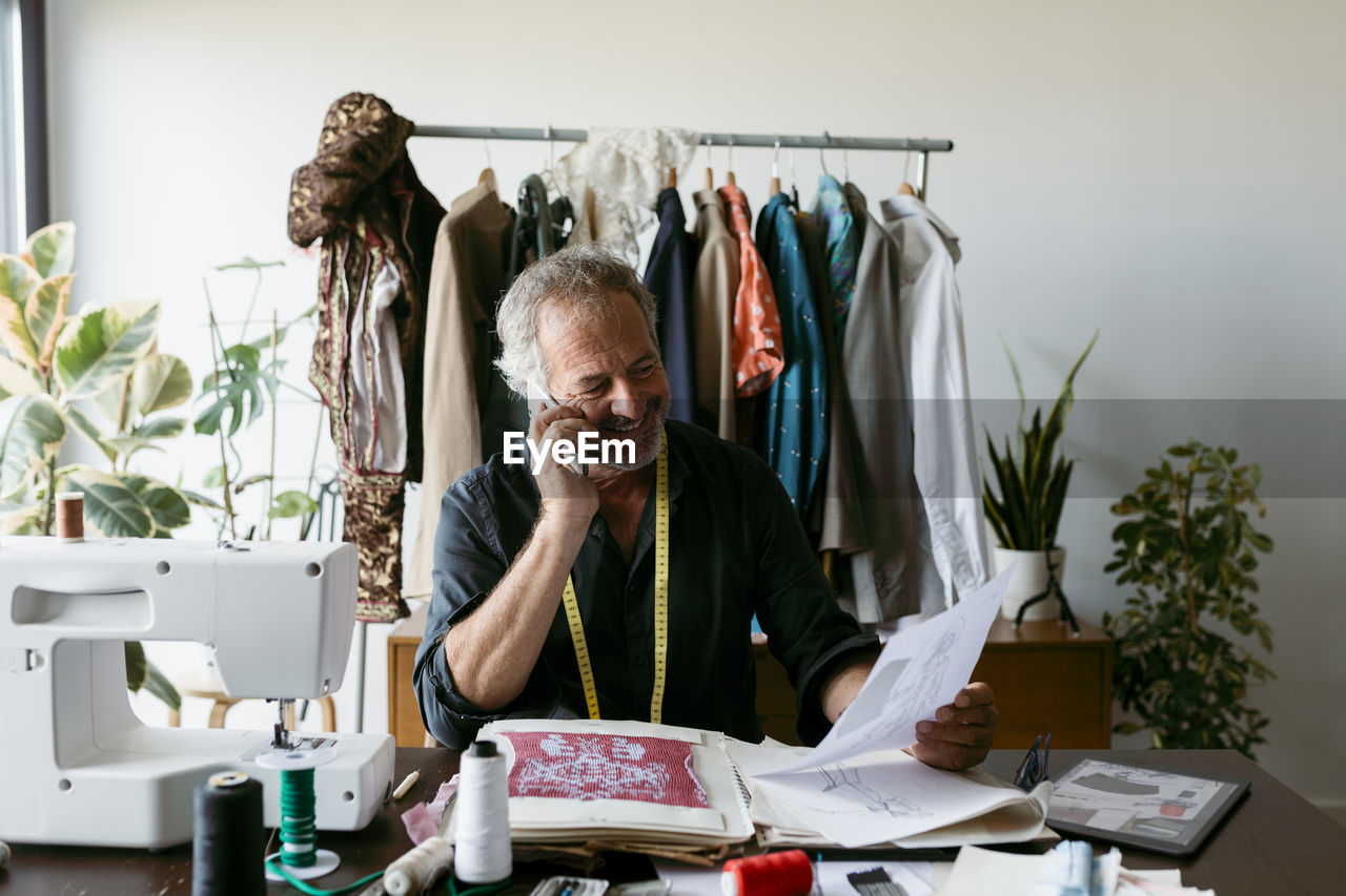 Smiling male tailor talking through smart phone while looking at design on paper