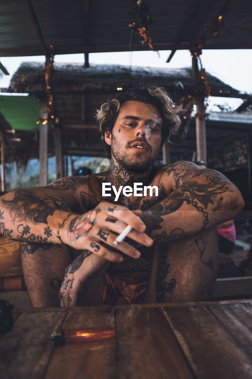 Portrait of shirtless man smoking cigarette sitting by table