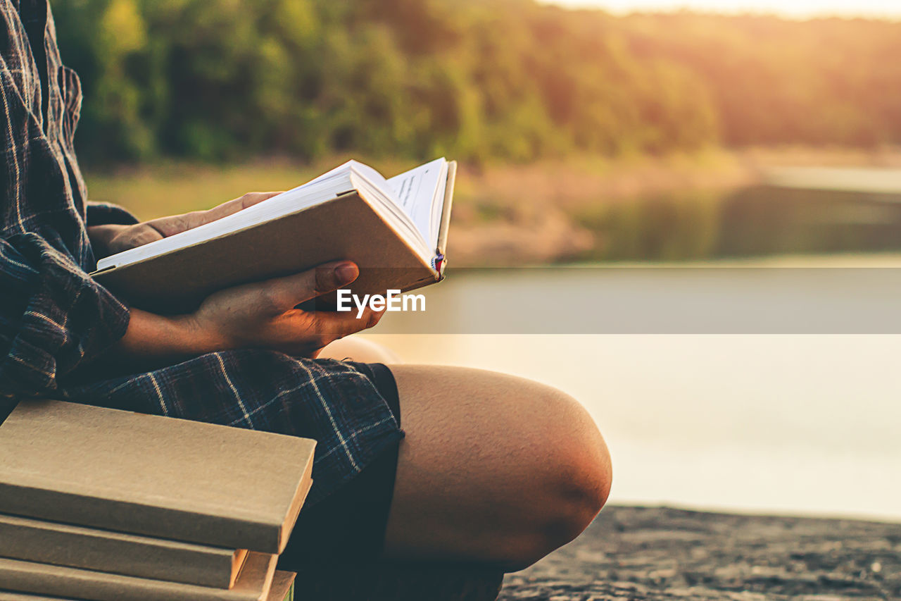 Midsection of woman reading book while sitting against lake