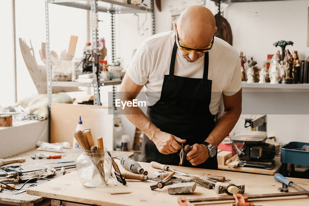 Focused mature woodworker choosing instruments from wooden workbench while working in creative workshop