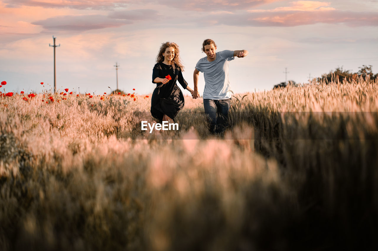 Happy young woman and man running on summer field against sky during sunset