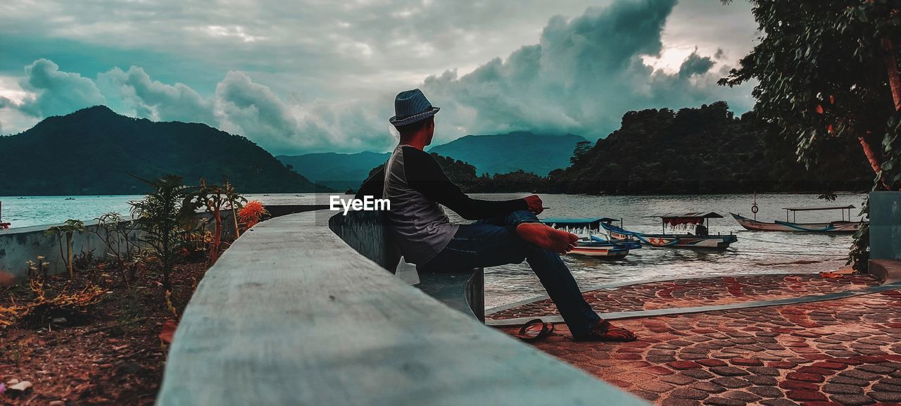 MAN SITTING ON BOAT IN SEA AGAINST MOUNTAINS