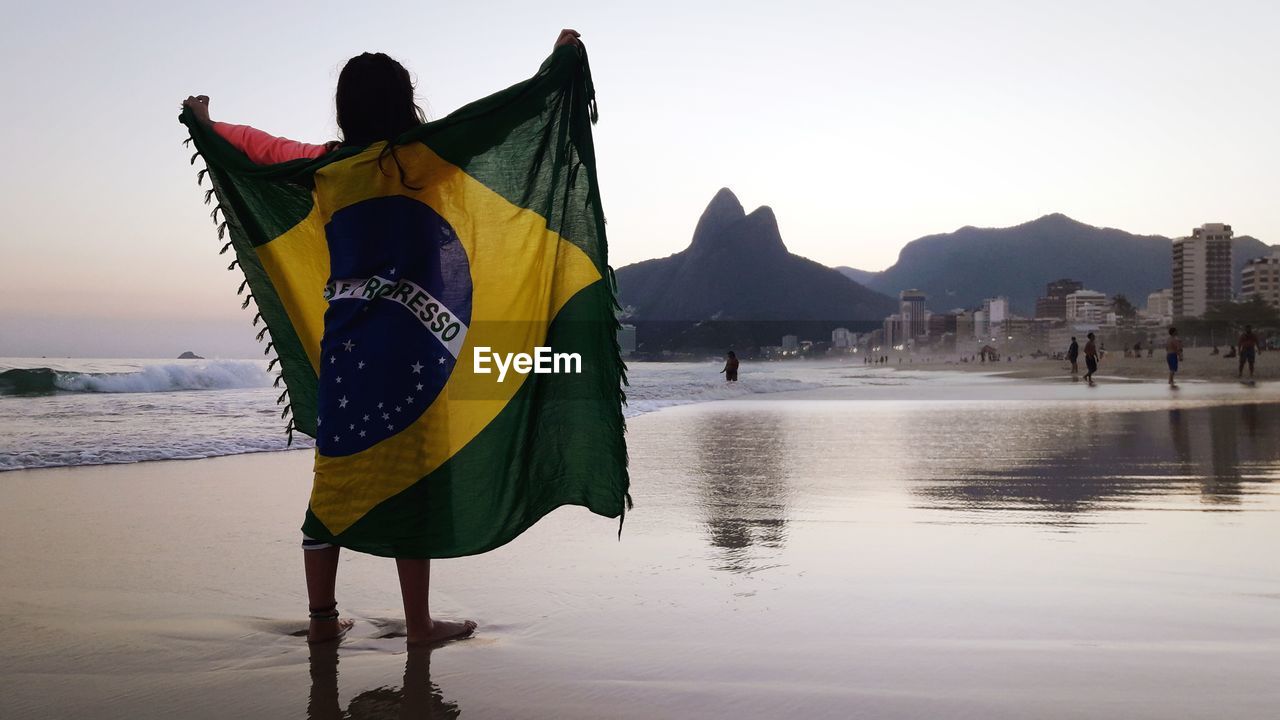 Rear view of girl holding brazilian flag while standing at beach against clear sky