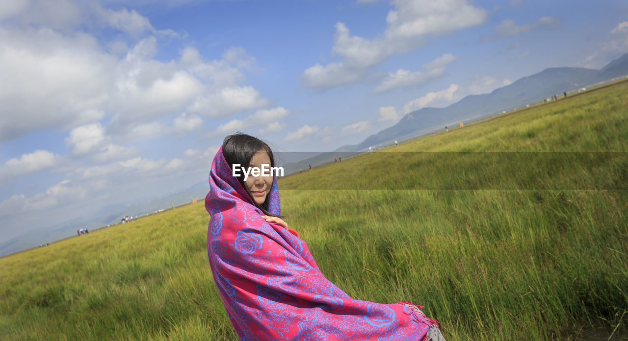 Portrait of woman wrapped in shawl sitting by grassy landscape against sky