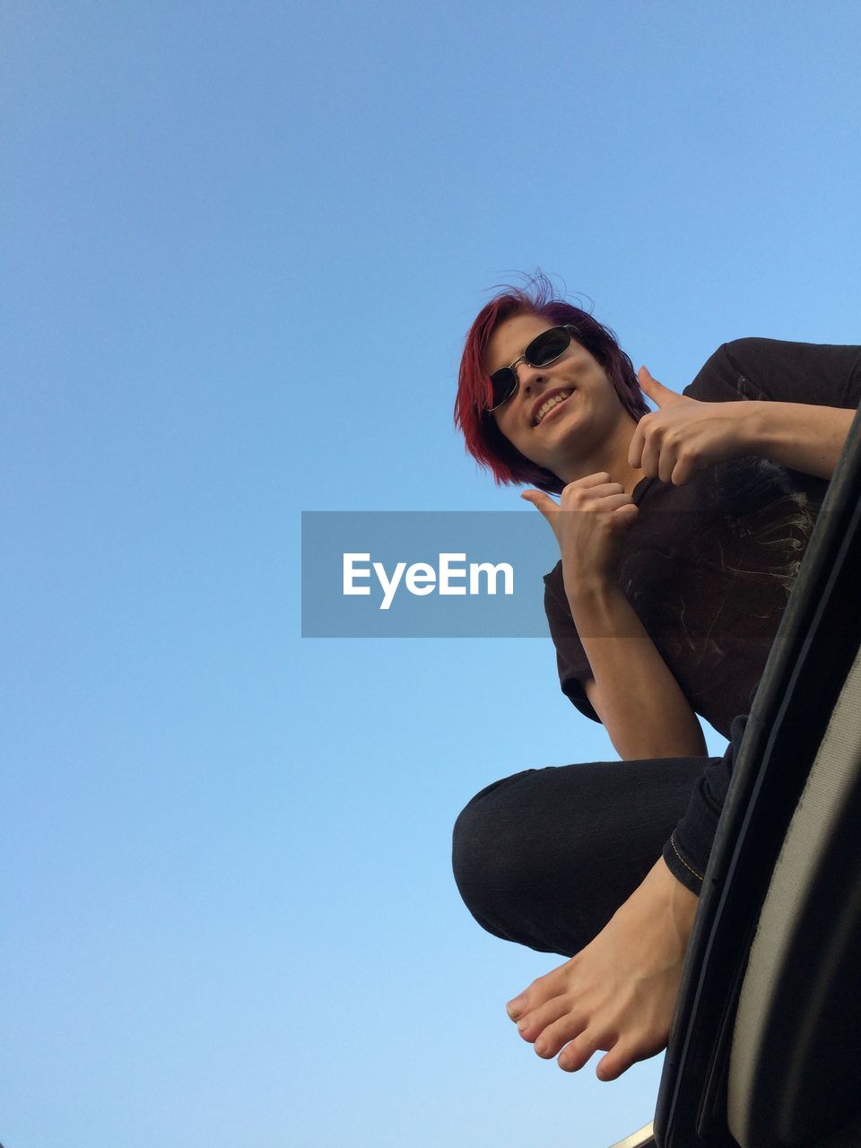 Low angle portrait smiling young woman showing thumbs up against clear sky