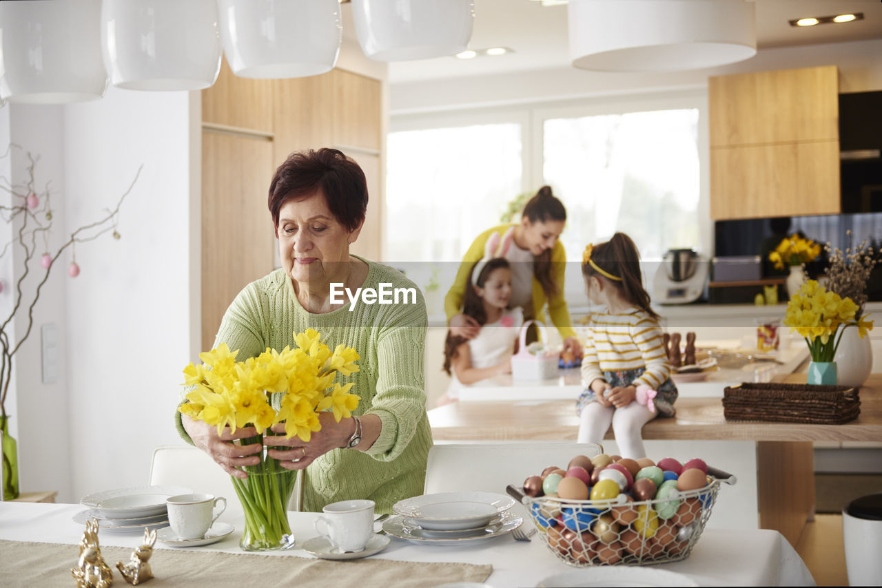 Senior woman arranging flowers on dining table with family in background