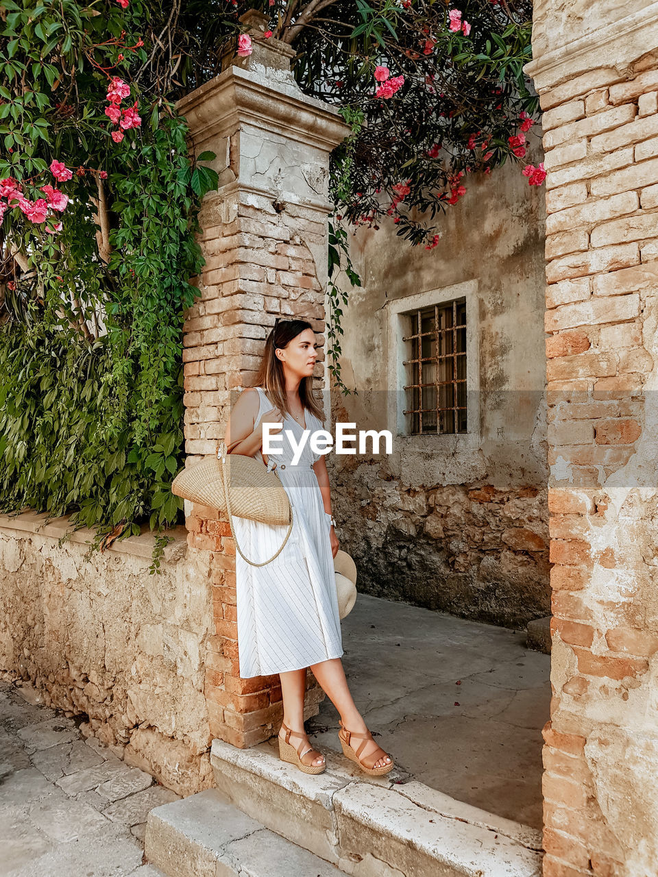 Full length portrait of woman in white summer dress leaning on brick wall.