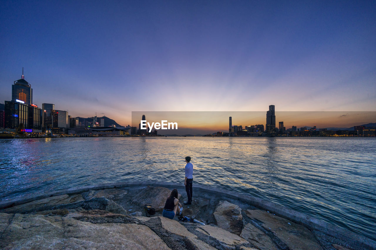 REAR VIEW OF MAN STANDING IN SEA AGAINST SKY DURING SUNSET