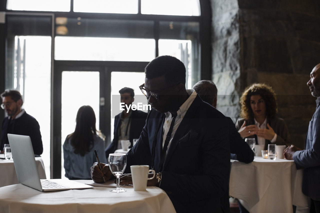 Businessman with laptop on table writing in notepad while standing at seminar