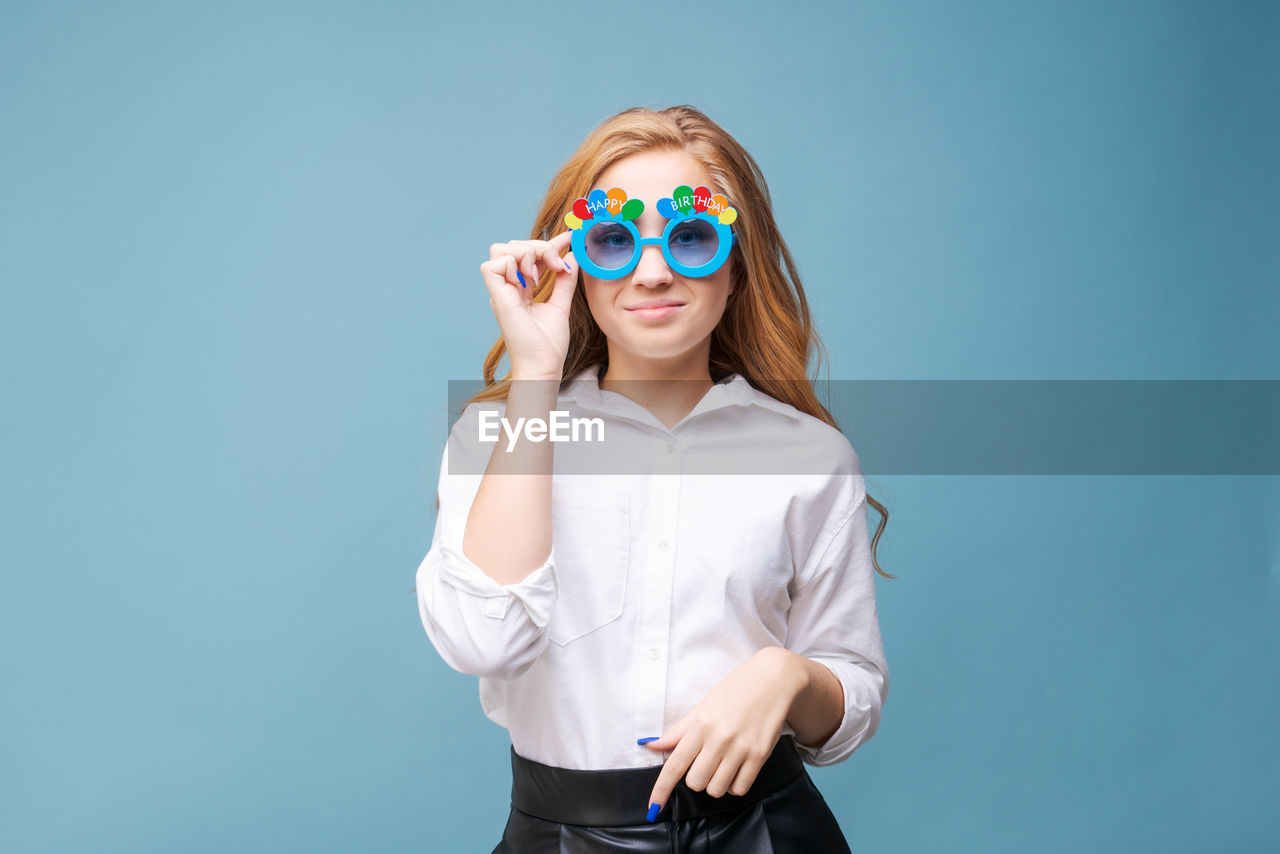 one person, studio shot, fashion, women, colored background, blue, glasses, adult, portrait, sunglasses, young adult, smiling, indoors, clothing, eyewear, happiness, fun, looking at camera, copy space, blue background, emotion, blond hair, female, hairstyle, front view, vision care, standing, goggles, waist up, human face, finger, photo shoot, long hair, cheerful, person, cute, humor, costume, positive emotion, child, cut out, lifestyles, childhood, casual clothing