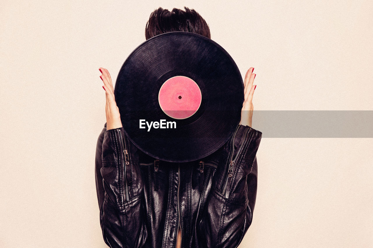 Faceless woman in leather jacket hiding face behind vinyl record in studio on pink background