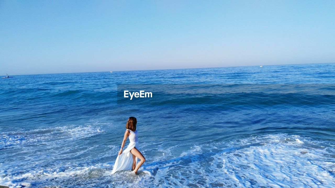 Side view of young woman standing in sea against clear blue sky