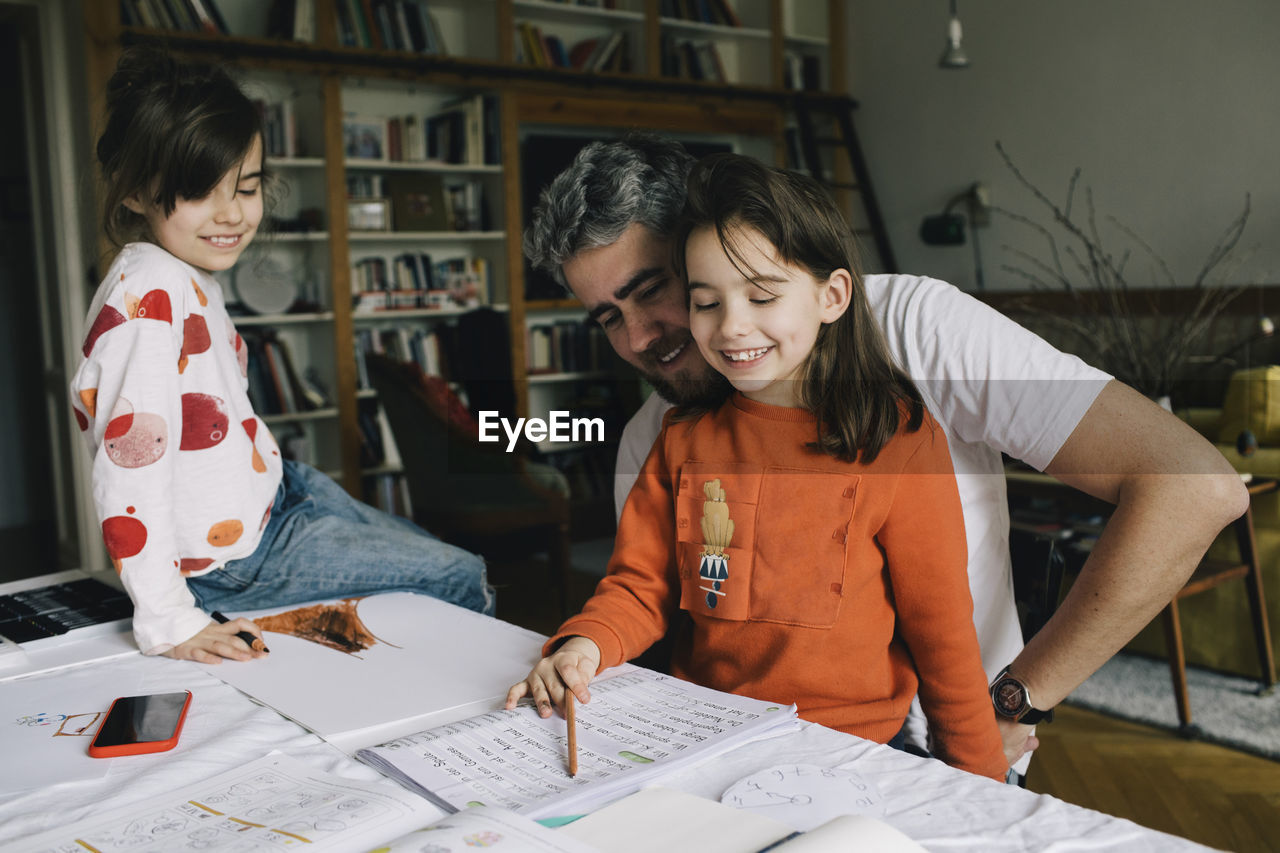 Smiling father looking at daughter reading book while girl sitting on table