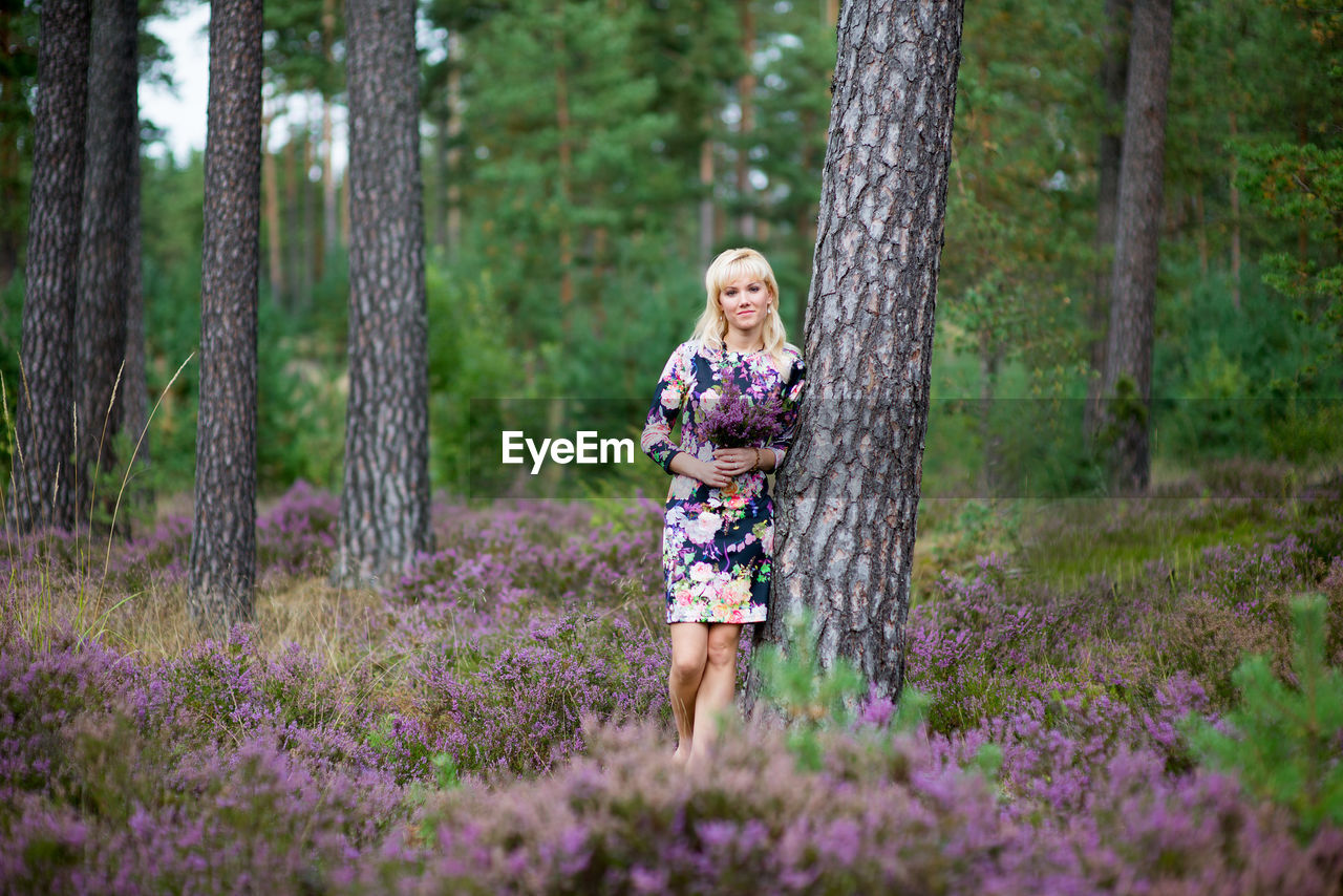 Woman standing by tree amidst purple flowering plants in forest