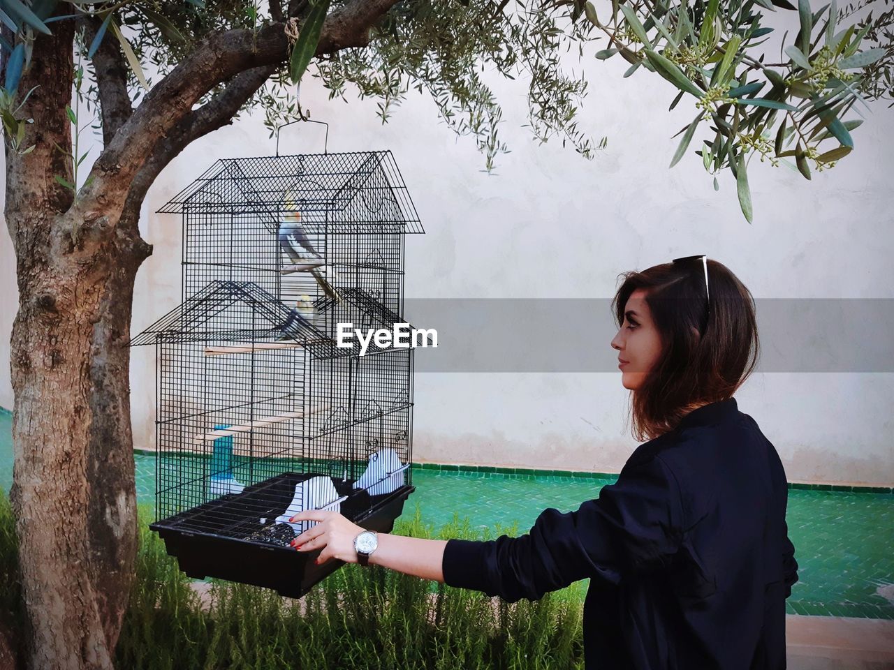 Side view of young woman looking at bird in cage while standing against wall