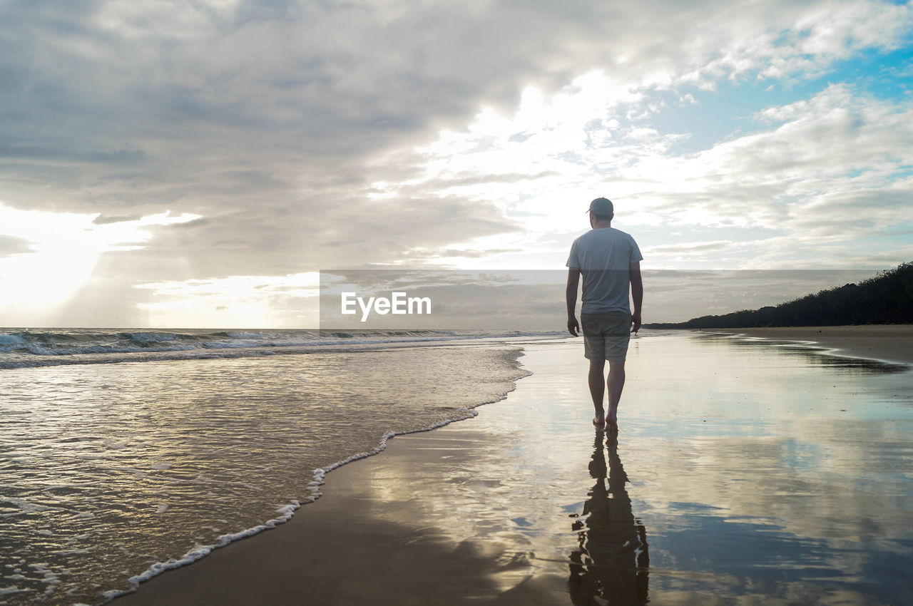 Rear view of man standing at beach against sky during sunset