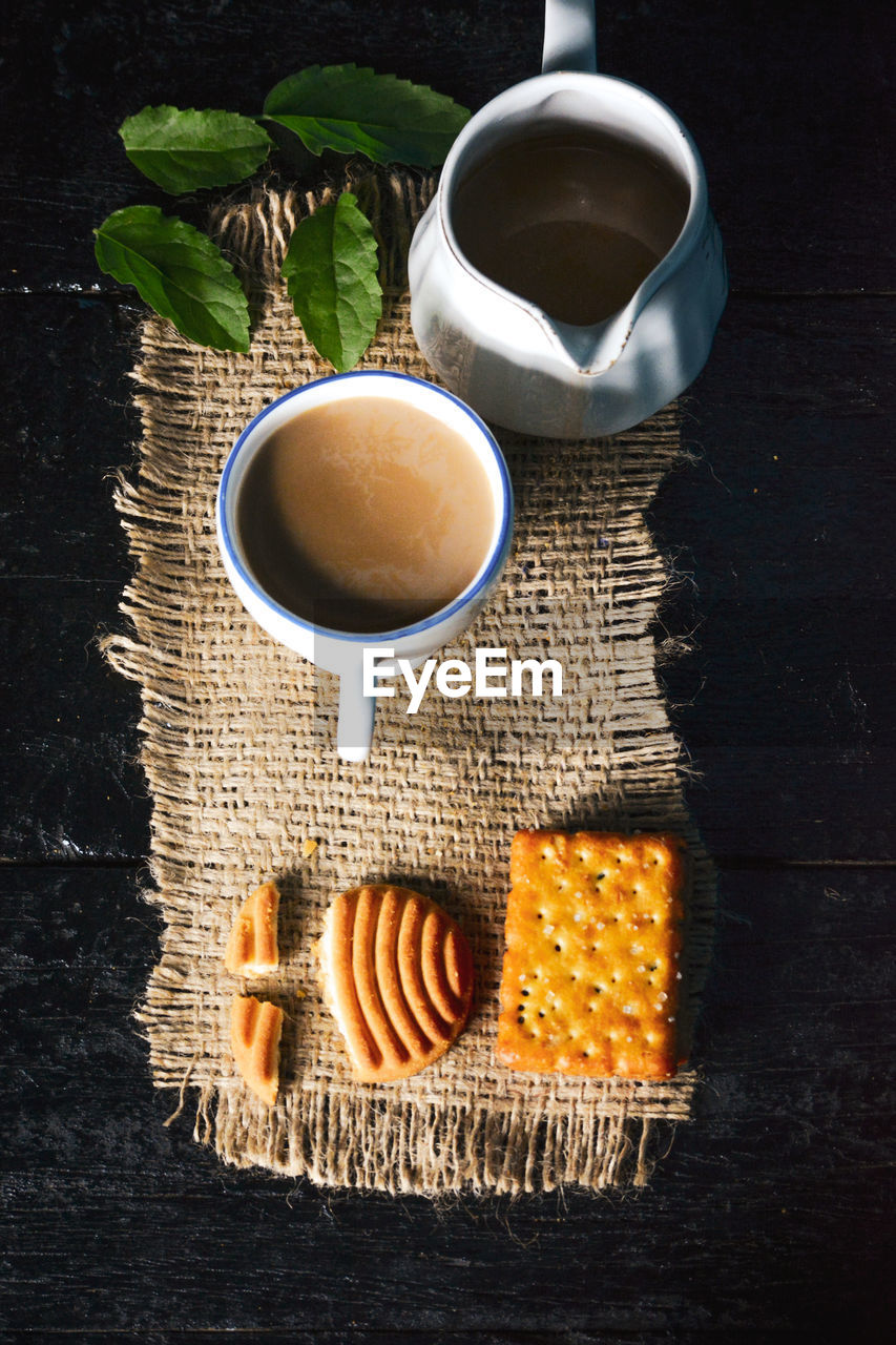 A cup of tea with tasty biscuits, teapot and fresh leaves on old wooden dark background