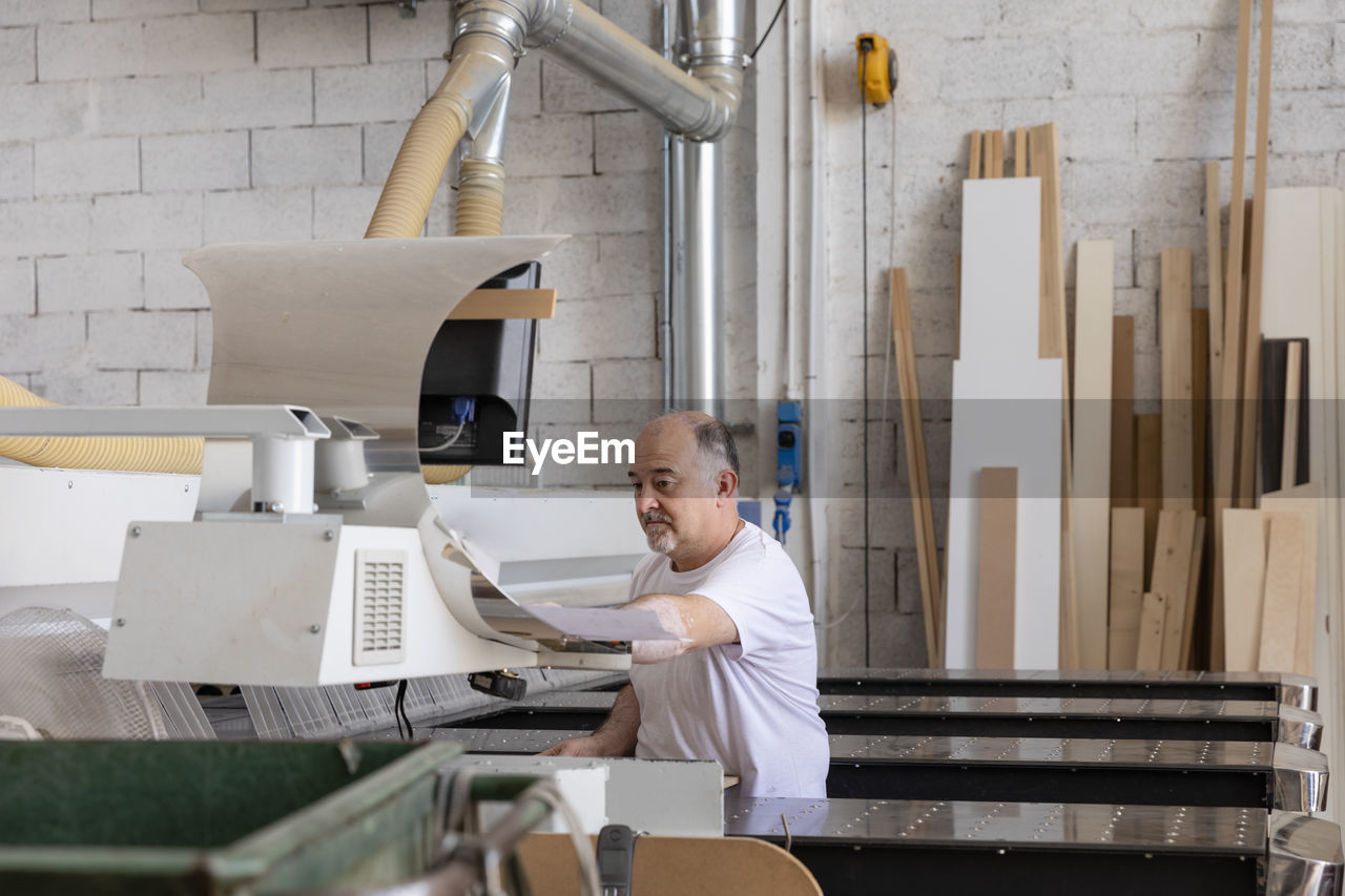 Senior man operating wood cutting machine while sitting at factory