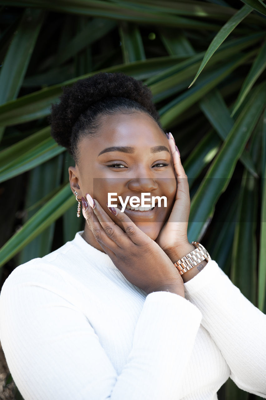 Positive african american female touching cheeks while standing near lush green plants on summer day in garden