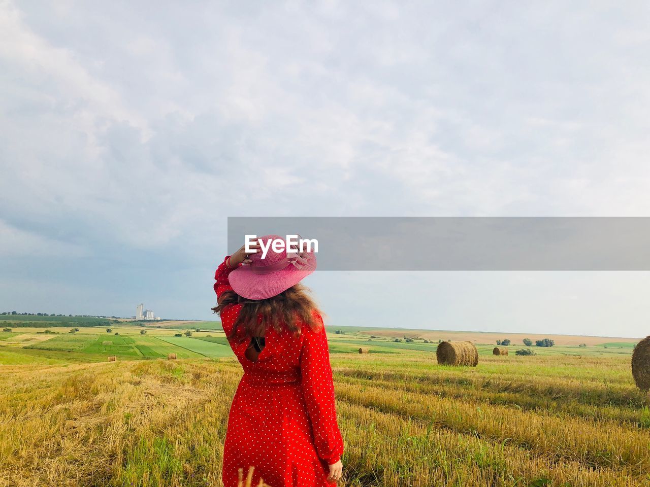 Back of woman wearing a red dress and a summer hat in the middle of a harvested field