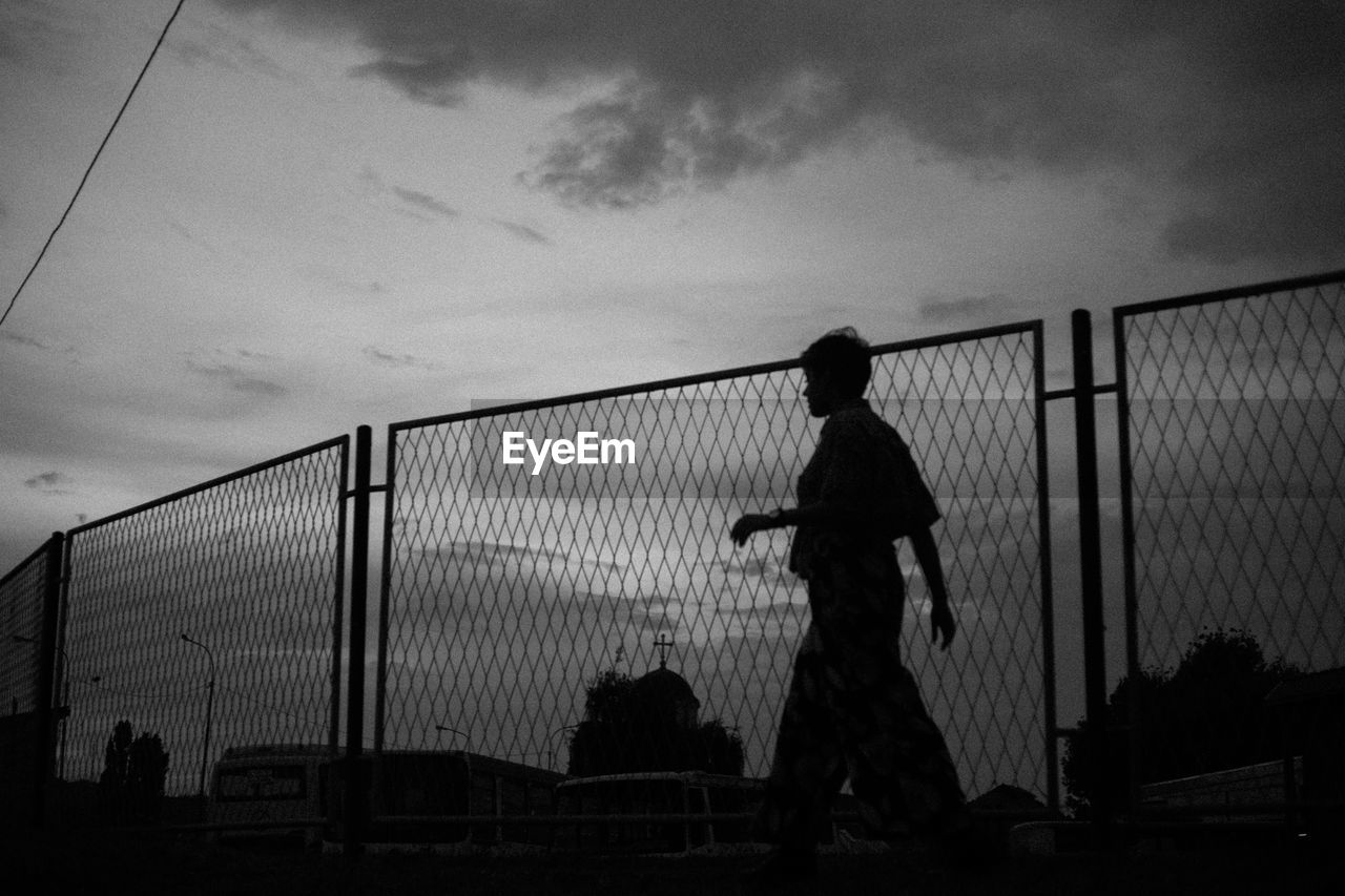 Silhouette woman standing by fence against sky during sunset