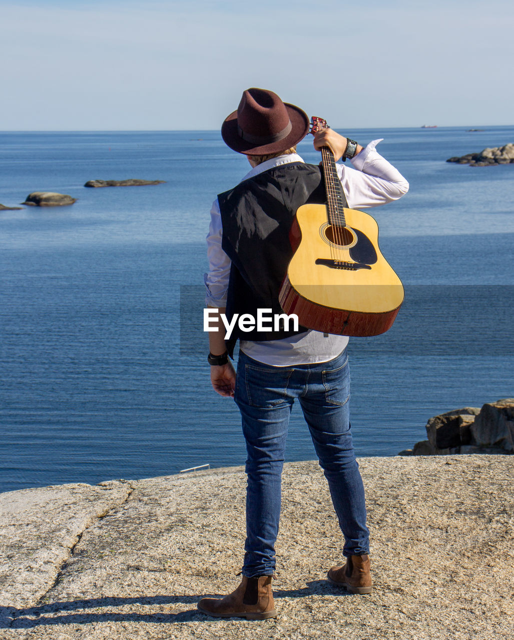 string instrument, water, sea, musical instrument, full length, beach, guitar, one person, music, blue, musician, casual clothing, standing, nature, hat, adult, land, coast, day, leisure activity, musical equipment, vacation, ocean, sky, men, sunlight, holding, clothing, arts culture and entertainment, rear view, shore, beauty in nature, person, outdoors, holiday, travel, trip, horizon over water, lifestyles, fashion accessory, young adult, violin, scenics - nature, jeans, plucking an instrument, sand