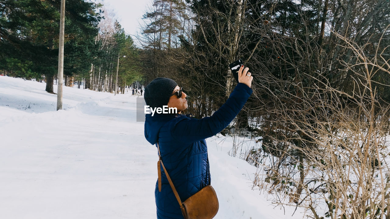 Side view of woman photographing with smart phone at forest during winter