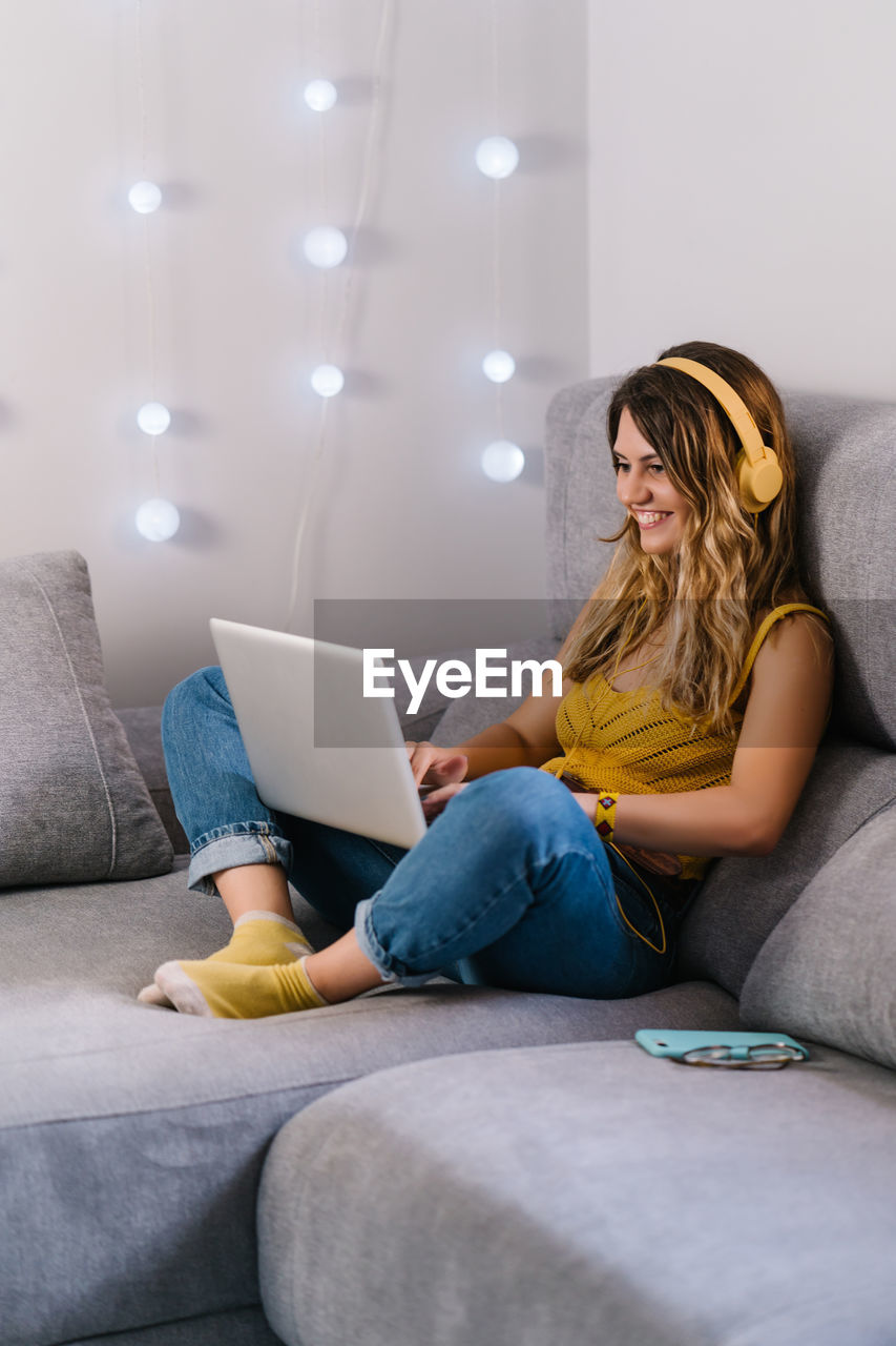 YOUNG WOMAN USING MOBILE PHONE WHILE SITTING ON SOFA