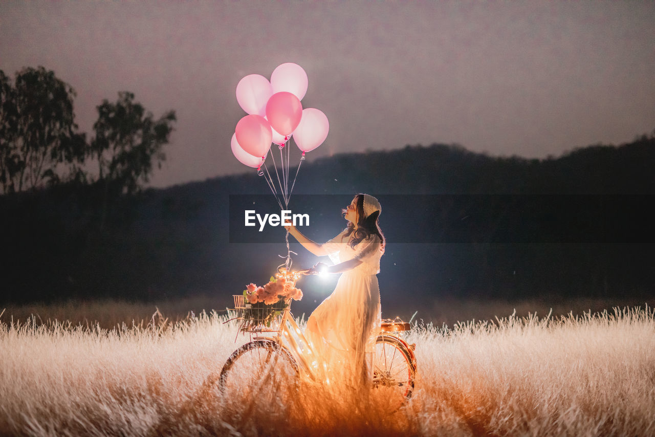 Woman with balloons sitting on illuminated bicycle on field against sky at sunset