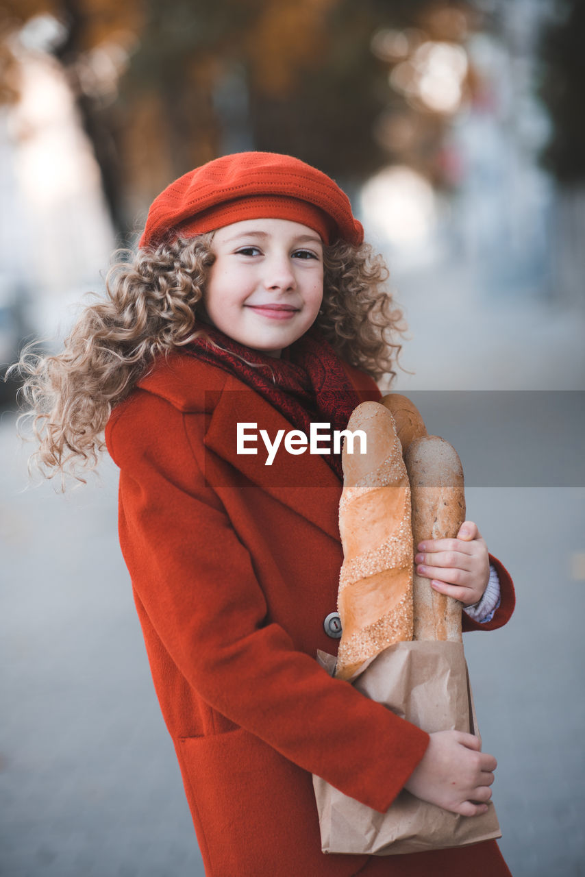 Smiling blonde happy child girl 5-6 year old with curly hair hold bread wearing hat and red jacket