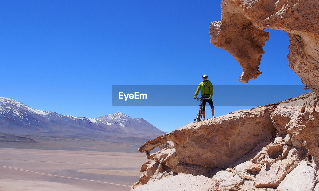 Man standing on rock with bicycle against blue sky