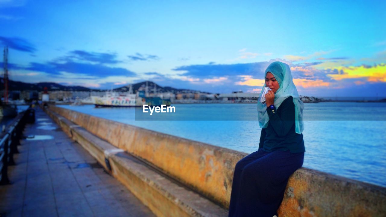 Thoughtful woman sitting on retaining wall by sea against sky during sunset