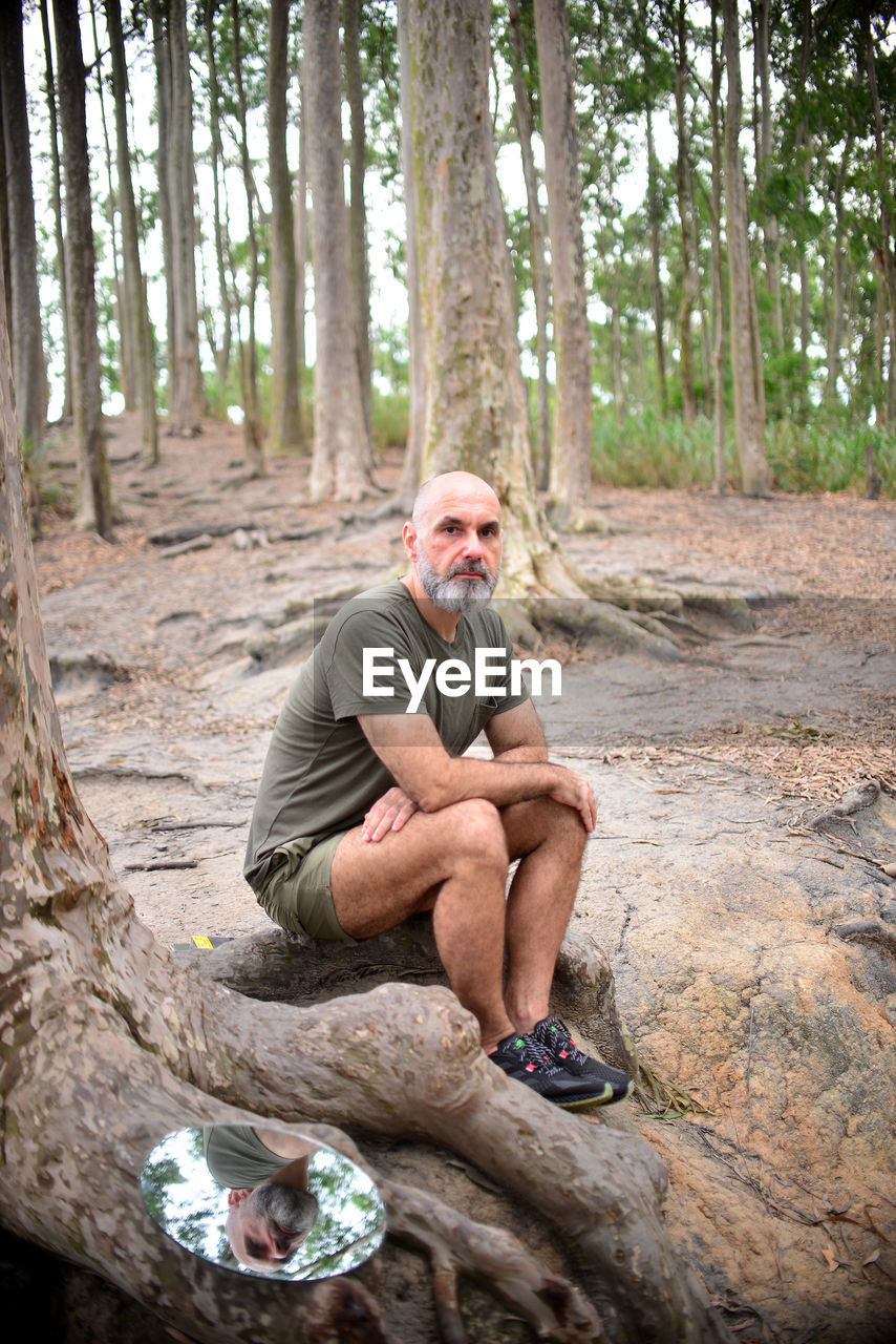 Full length portrait of man sitting by tree trunk in forest