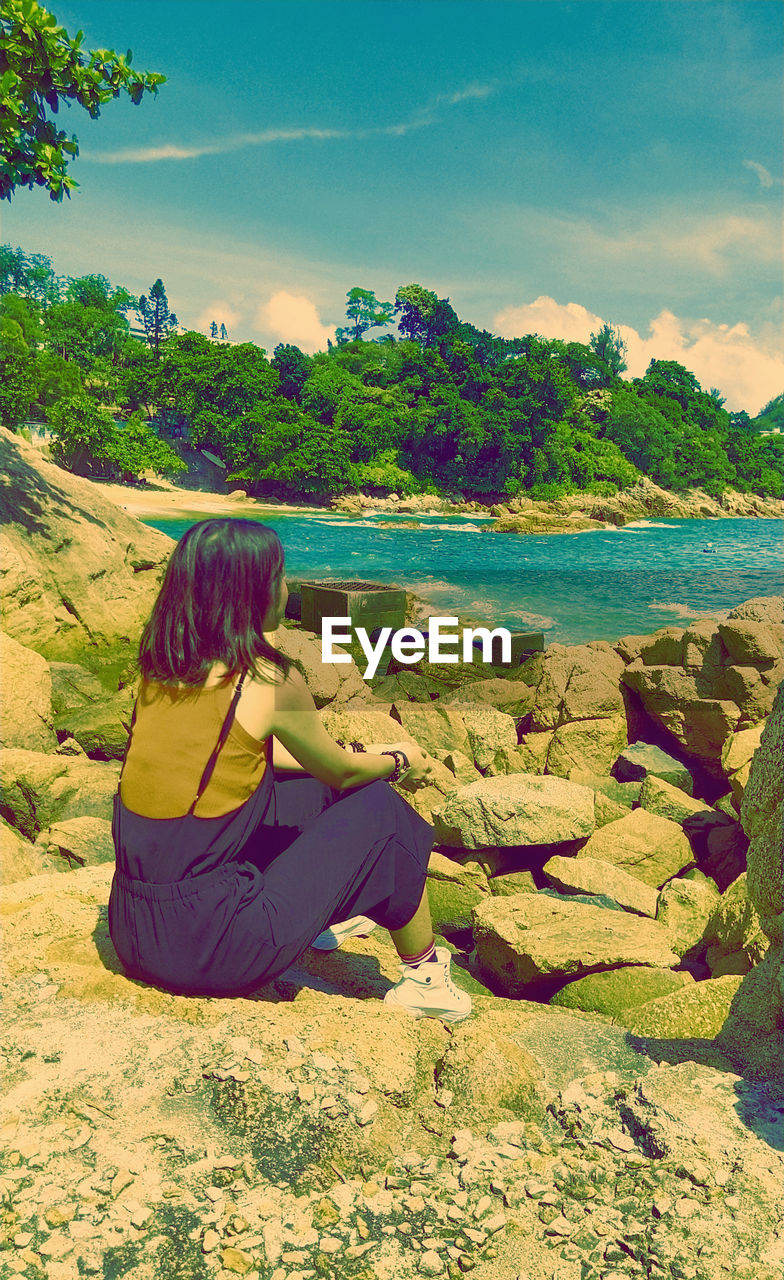 REAR VIEW OF WOMAN SITTING ON SHORE AT BEACH AGAINST SKY