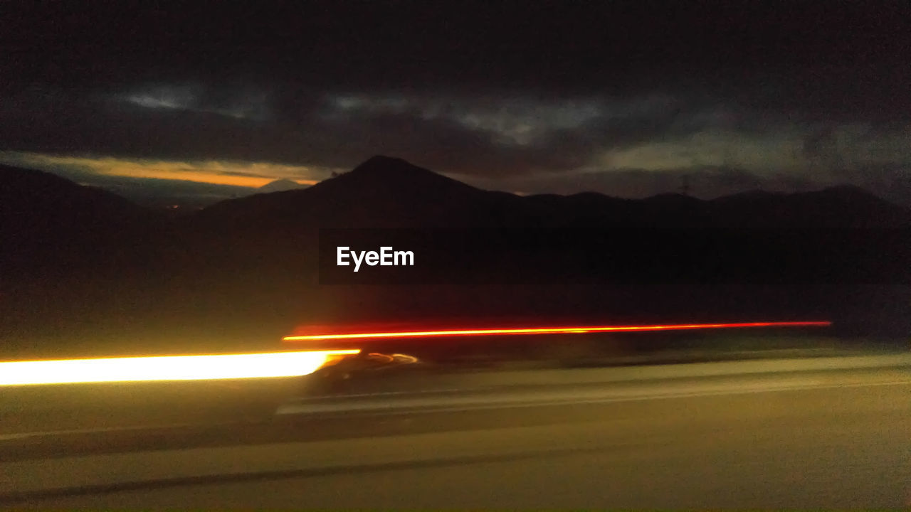 LIGHT TRAILS ON ROAD AGAINST MOUNTAIN AT NIGHT