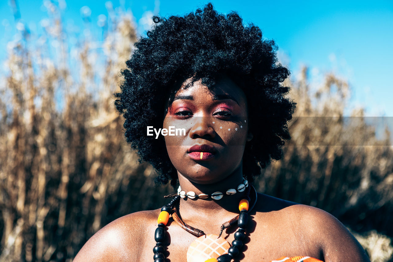 Serious african model with afro hairstyle wearing colorful handwoven kente and looking at camera while standing on field on bright summer day