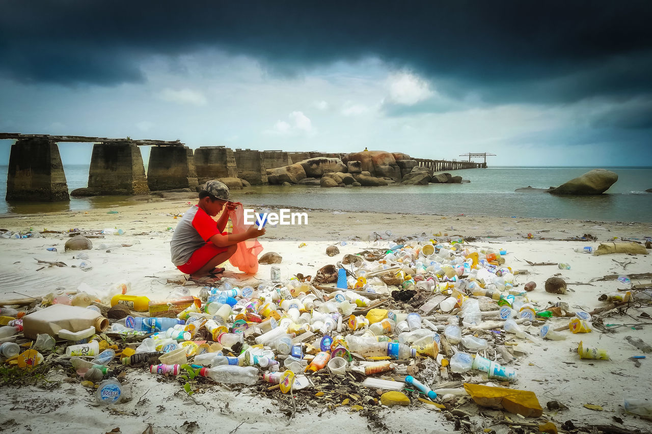 Side view of boy cleaning garbage while crouching at beach against cloudy sky