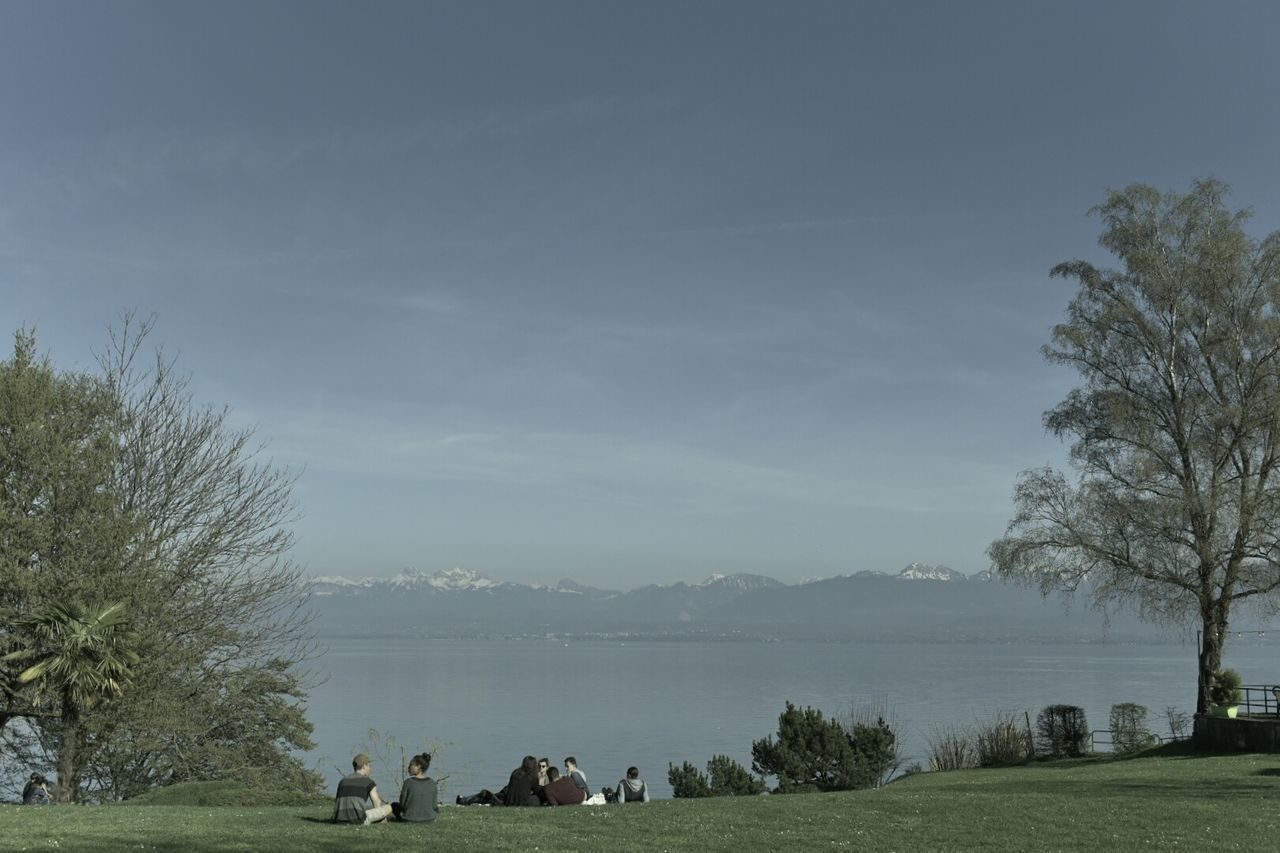 People sitting on grassy field by river against sky on sunny day