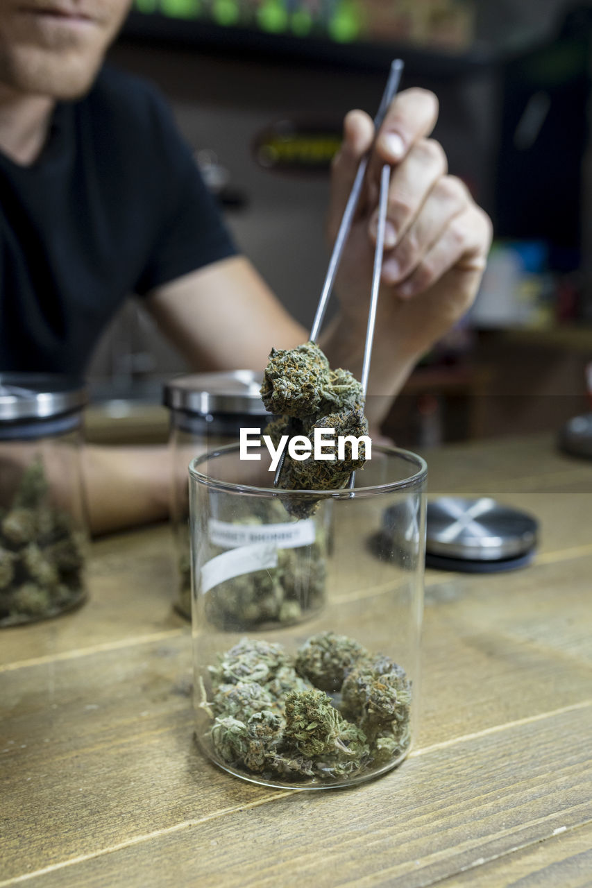 Crop anonymous male with tweezers taking out dry cannabis flower buds of jar on table in workspace