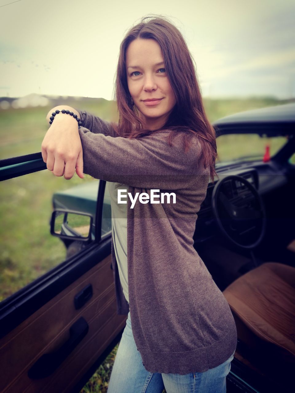 PORTRAIT OF SMILING YOUNG WOMAN SITTING ON CAR