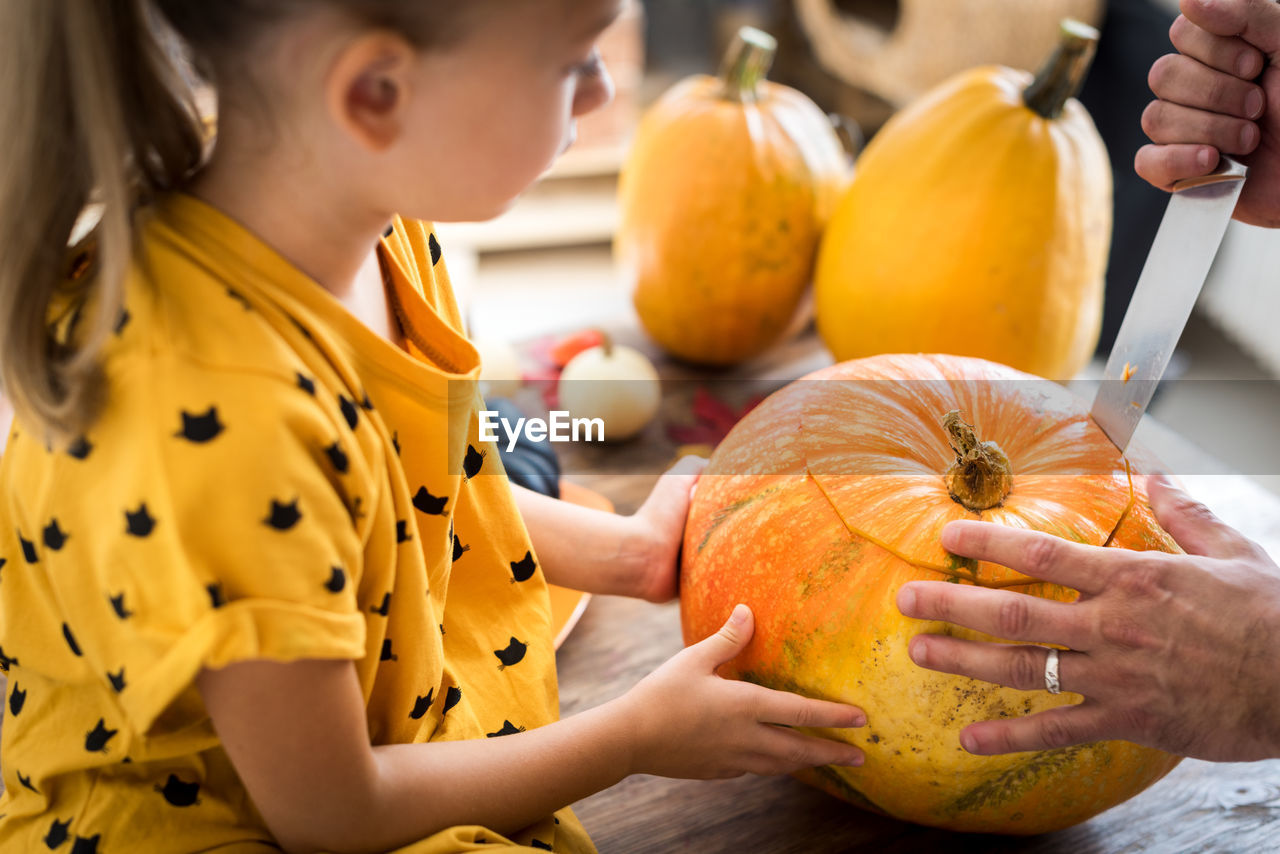 Cropped hand cutting pumpkin with girl sitting a home