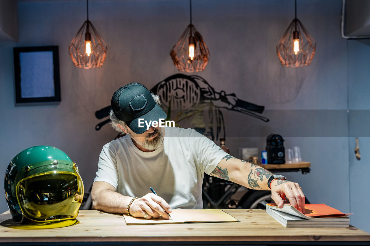 Front view of a man writing in a notebook on a wooden table next to a motorcycle helmet person