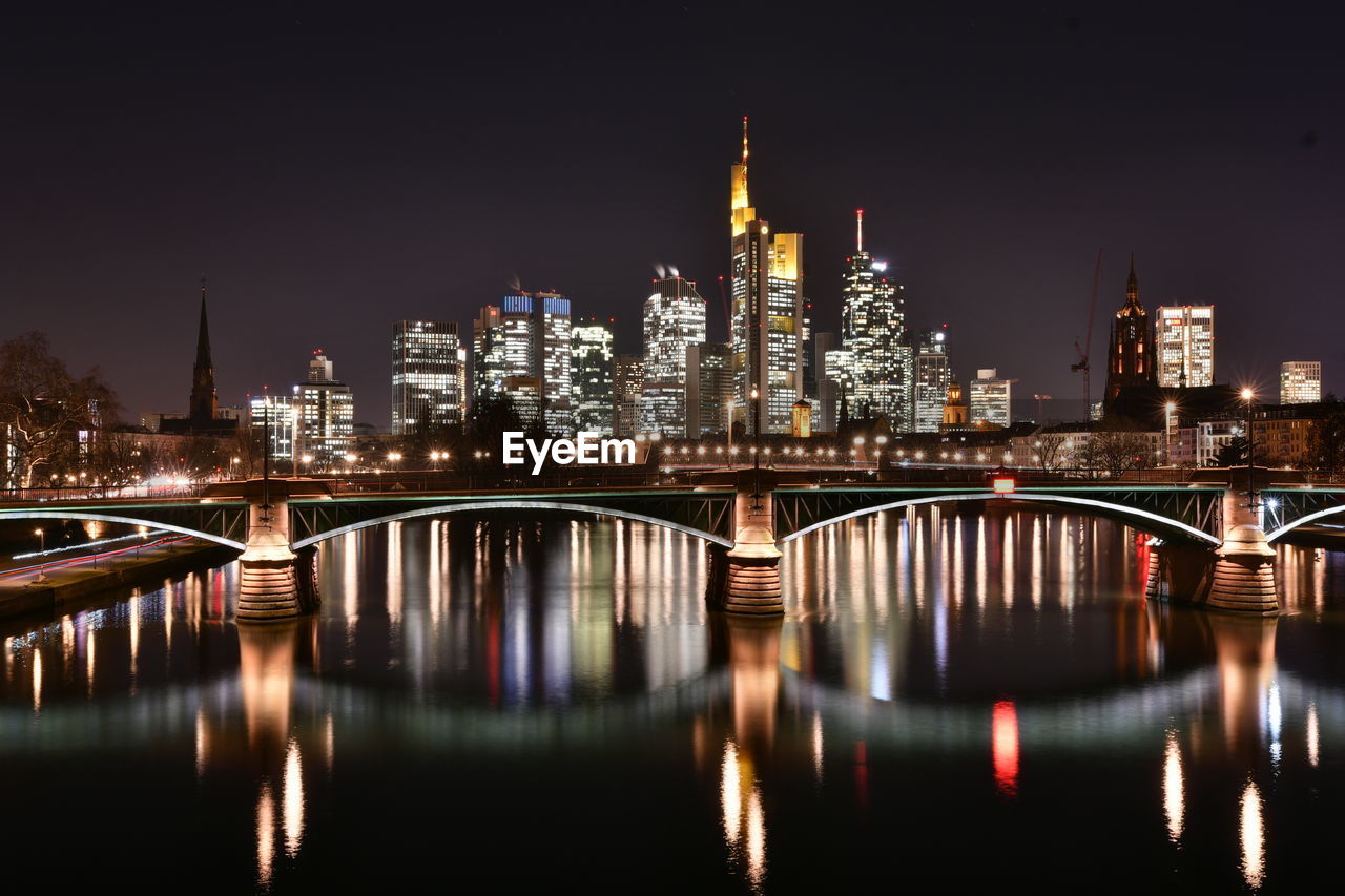 ILLUMINATED BRIDGE OVER RIVER BY BUILDINGS IN CITY AT NIGHT