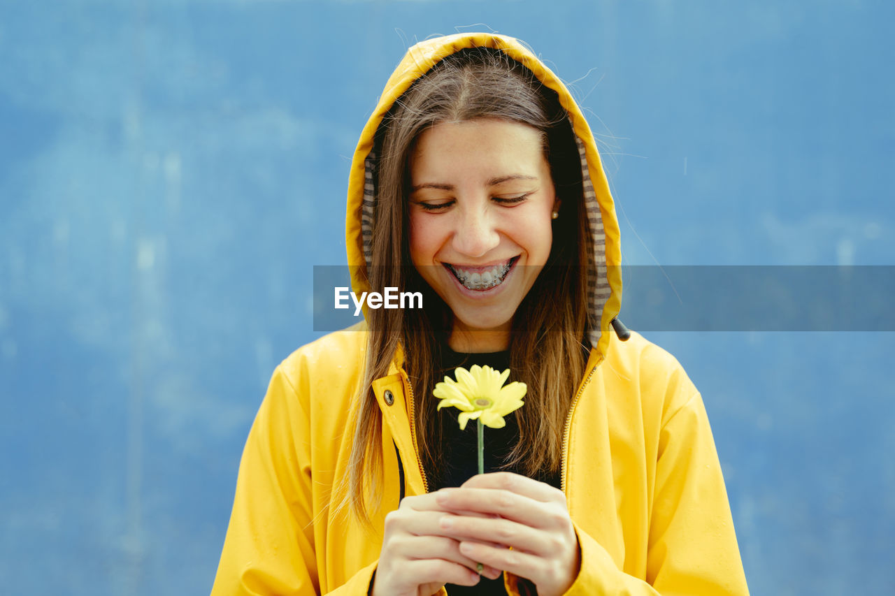 Smiling woman looking at flower standing against blue background