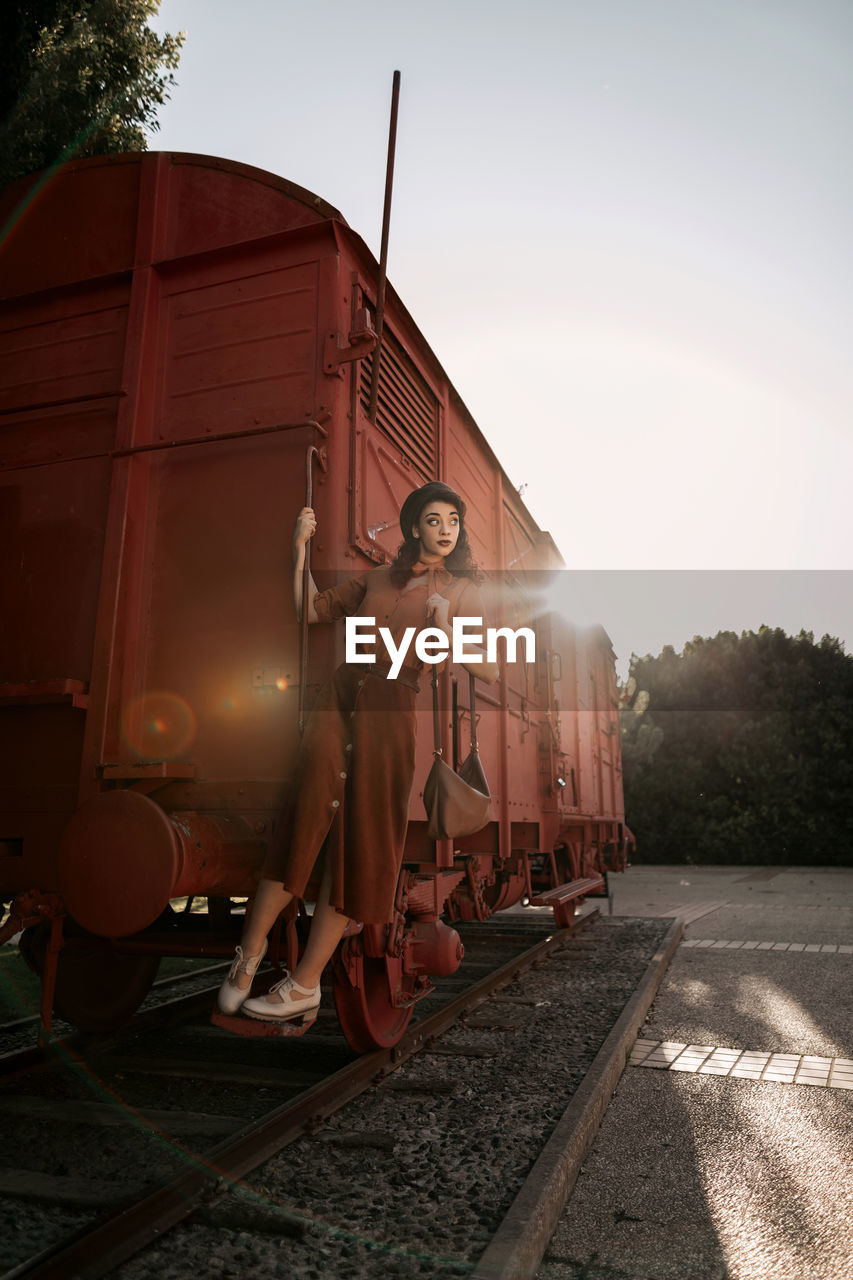 Young female with dark curly hair in beret wearing terracotta clothes in vintage style in back lit standing on step of car train painted in terracotta color and holding handrail while looking away flirty