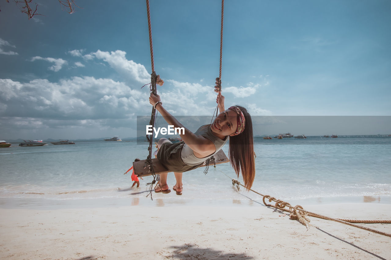 Portrait of smiling young woman sitting on swing at beach against sky