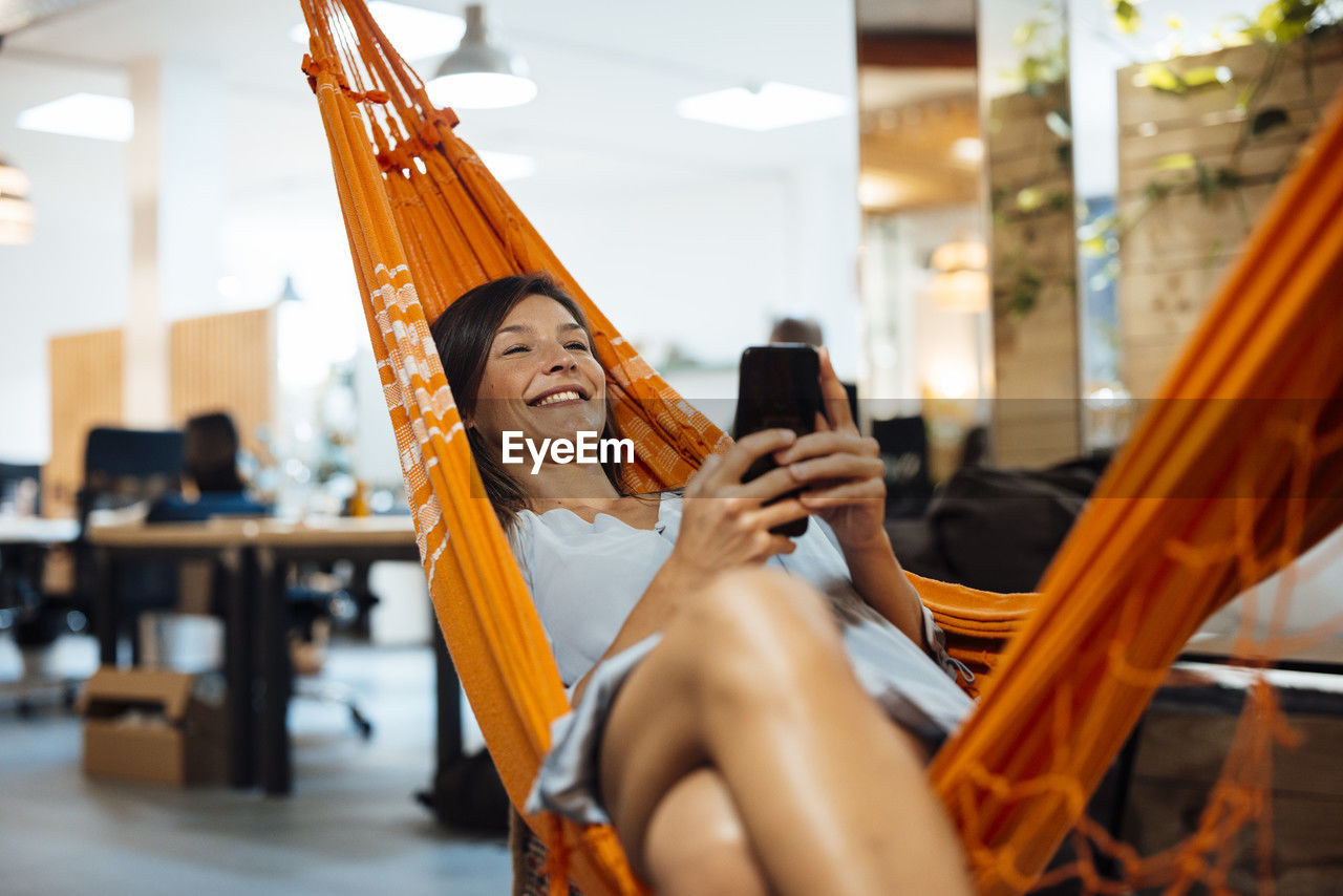 Smiling businesswoman using smart phone relaxing in hammock in office